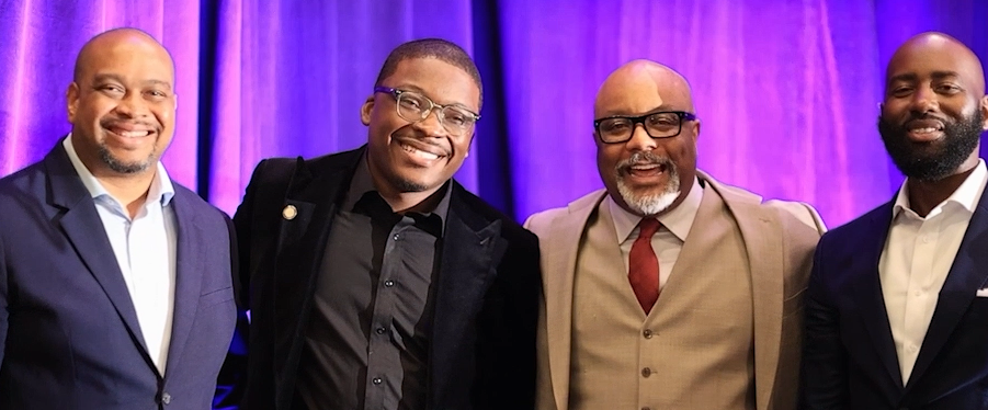 Four men in suits posing together in front of a purple backdrop