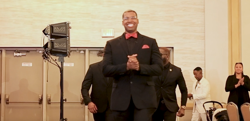 Man in a black suit and red bowtie smiling as he enters the ballroom