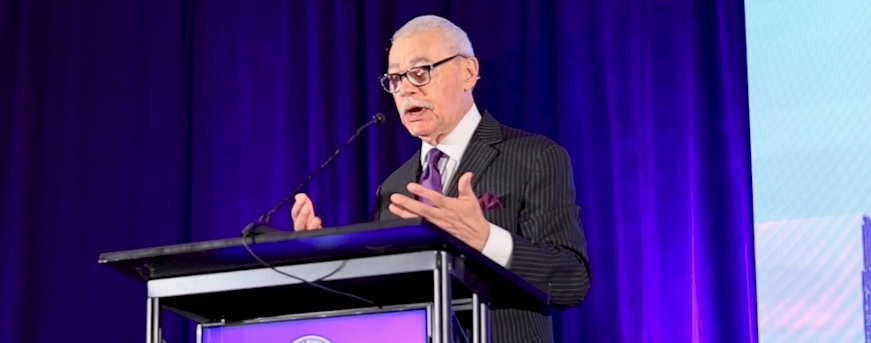 Distinguished speaker delivering remarks at the podium under purple stage lights